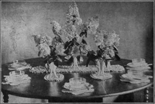A table decoration for a luncheon party. Two shades of mauve lilac are arranged in white china vases. The mats on which the vases and finger bowls stand are of crepe paper to match the lilac