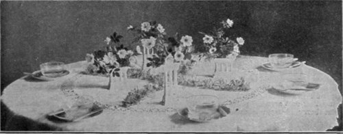 A pretty suggestion for a wild flower table decoration. Wild roses are arranged in white china column vases and separated by thick clusters of honeysuckle laid crosswise on the cloth. If candlesticks are used, they should be of white china